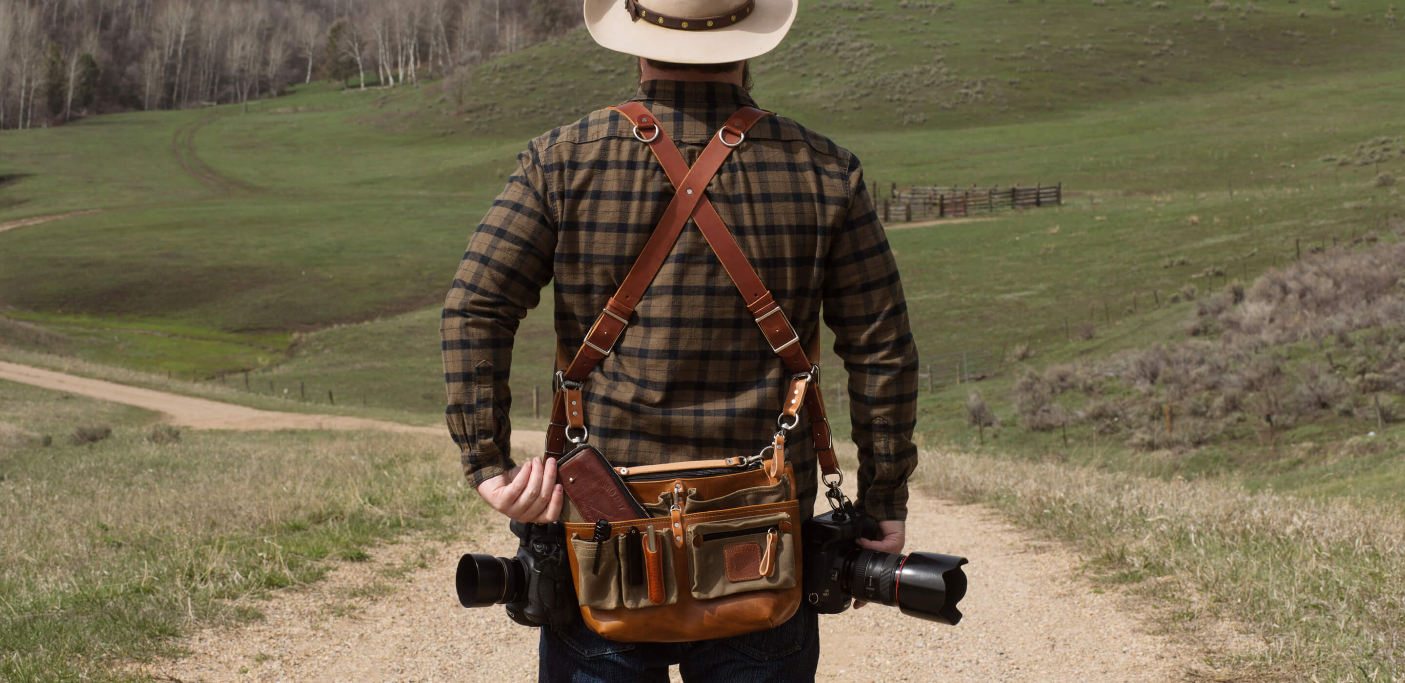 Person with a camera and leather bag walking on a dirt path in a scenic landscape.